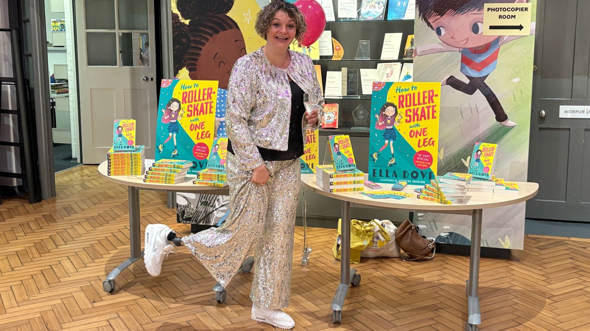 Ella Dove at her book launch, standing on one leg with her prosthetic leg kicked out to the side