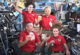 four astronauts float weightless in a cramped laboratory while holding plastic bags of freeze-dried foods