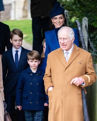 King Charles wearing a tan coat standing next to Prince Louis while leaving church on Christmas Day 2023, with Prince George and Princess Kate standing behind them