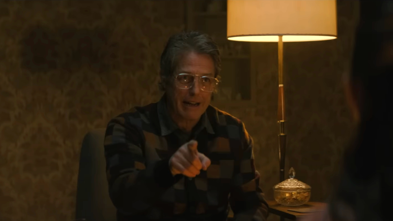 Hugh Grant señala hacia adelante con una mirada de humor mientras se sienta en la silla de su sala en Heretic.