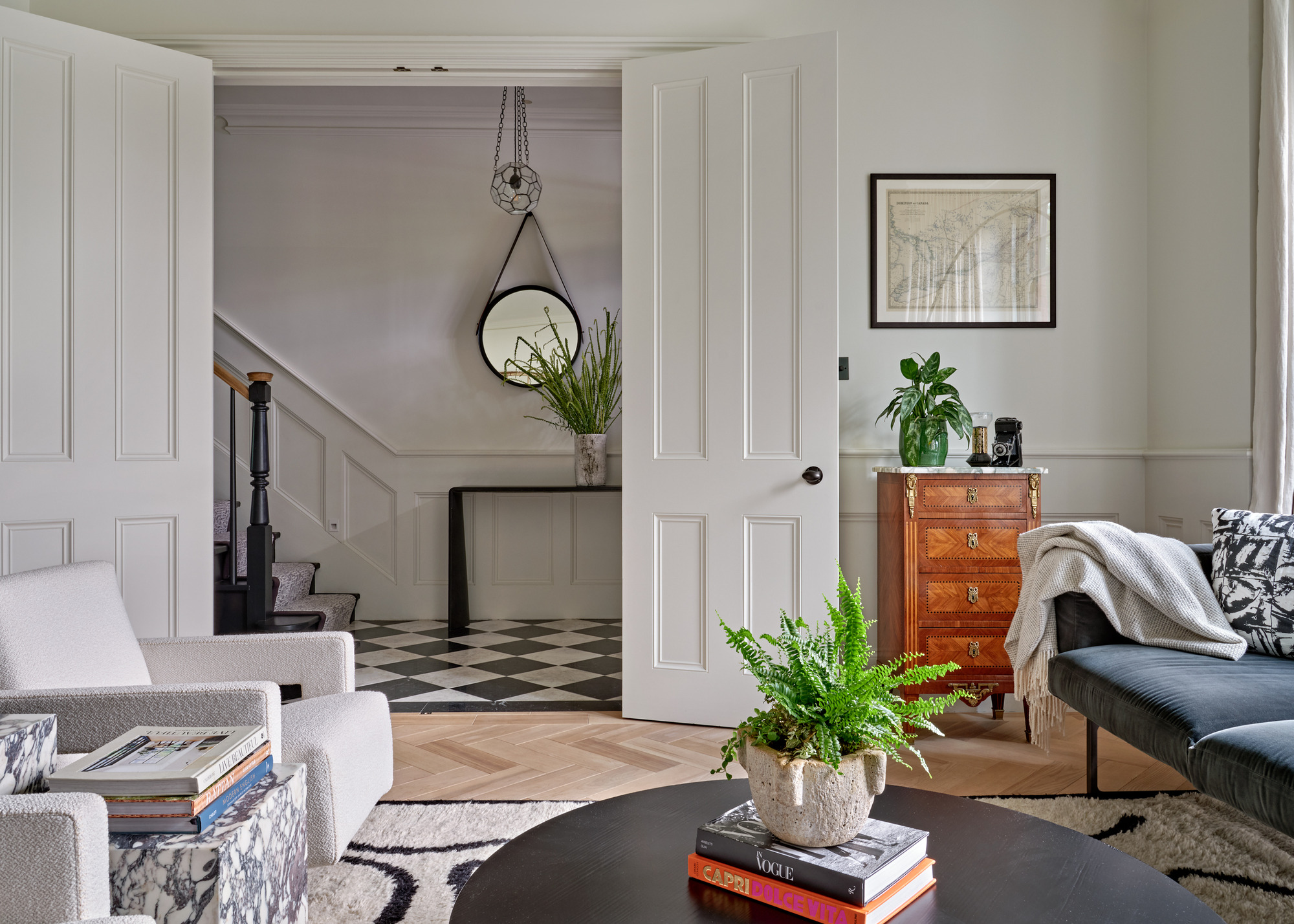 A white living room with wide double doors leading to a hallway with a checkered floor; there's a dark sofa, two cream armchairs and a black round coffee table in the center