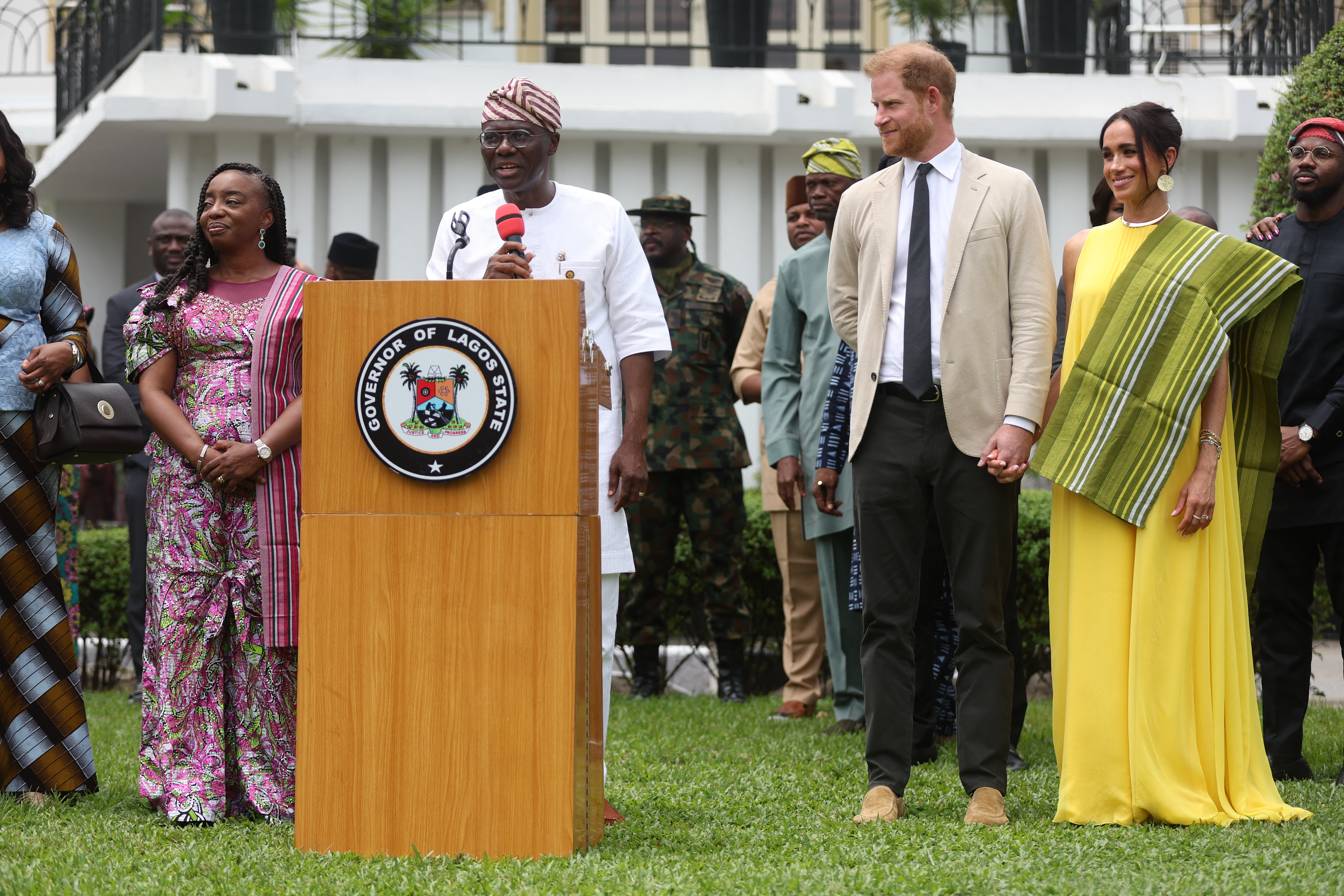 Prince Harry and Meghan Markle standing next to a man speaking at a podium in Nigeria