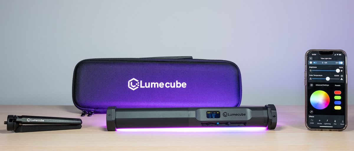 Lume Cube RGB Tube Light Mini emitting a purple light, in front of the Lume Cube case, tripod (left) and a phone displaying the Lume Control app 