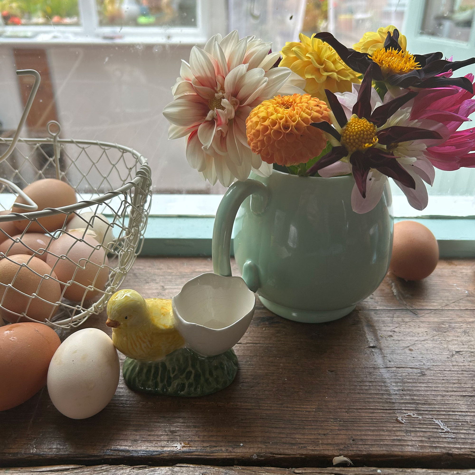 Small blue jug filled with dahlias beside wire basket filled with fresh eggs