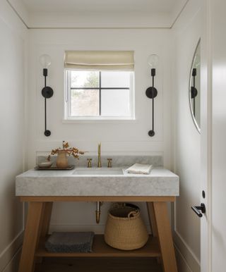 white powder room with marble basin on a wooden vanity, black wall lights, blind, brass taps