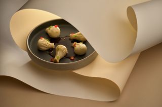 Five beautifully presented meat dumplings lay in a black dish surrounded by a cream-shaded swathe of paper arranged in a curvilinear composition.