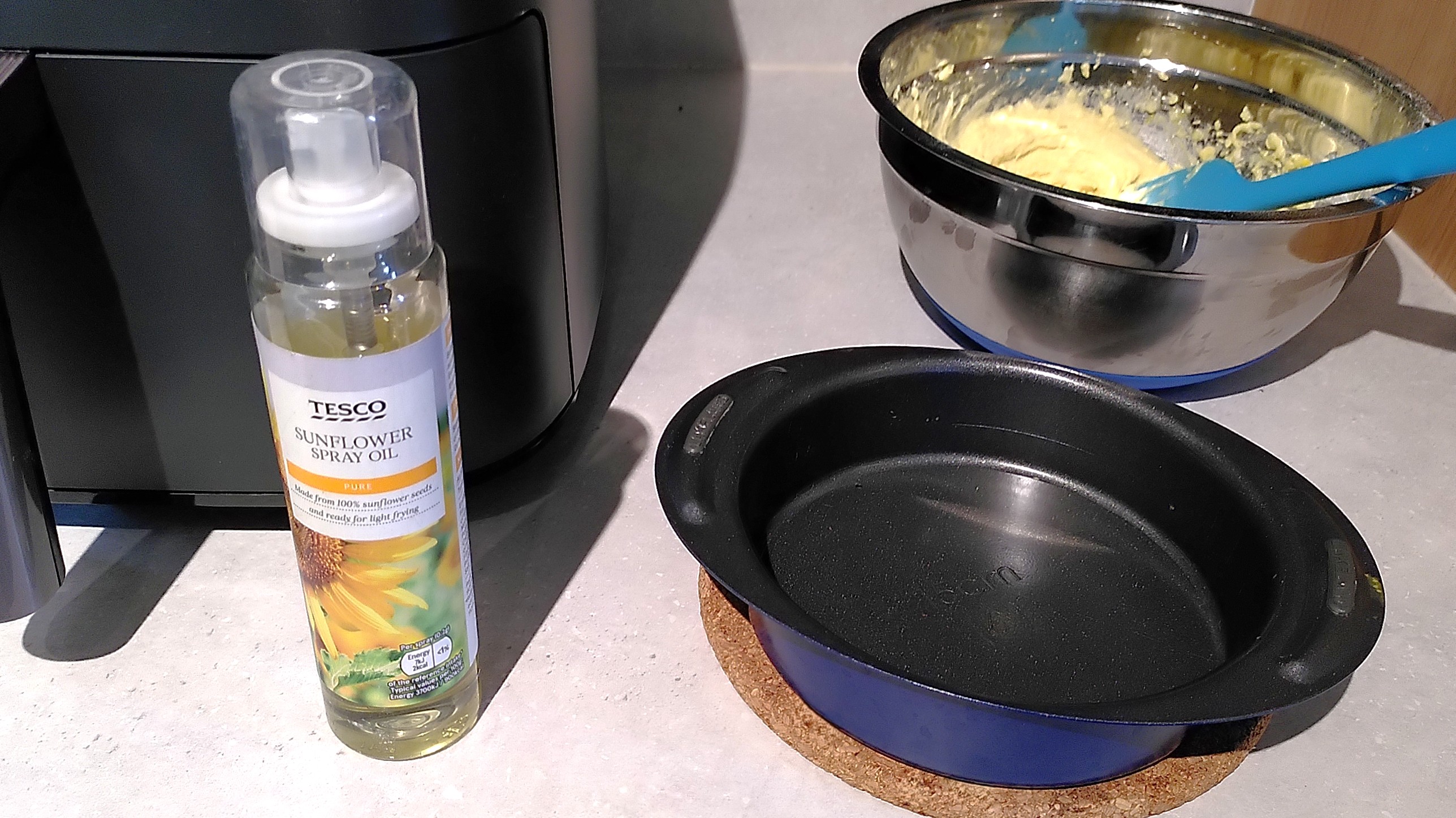 Air fryer with bowl of cake batter, cake tin, and sunflower oil spray