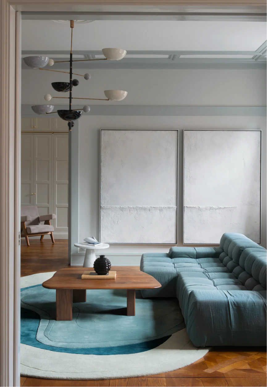 Modern living room with a teal sectional sofa, wooden coffee table, sculptural chandelier, and an irregular layered Erik Bratsberg Lozza Wool rug in soft blue tones anchoring the space.