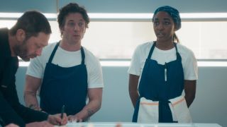 From left to right: Richie leaning into the frame, Carmy standing behind a counter and Sydney standing next to him.