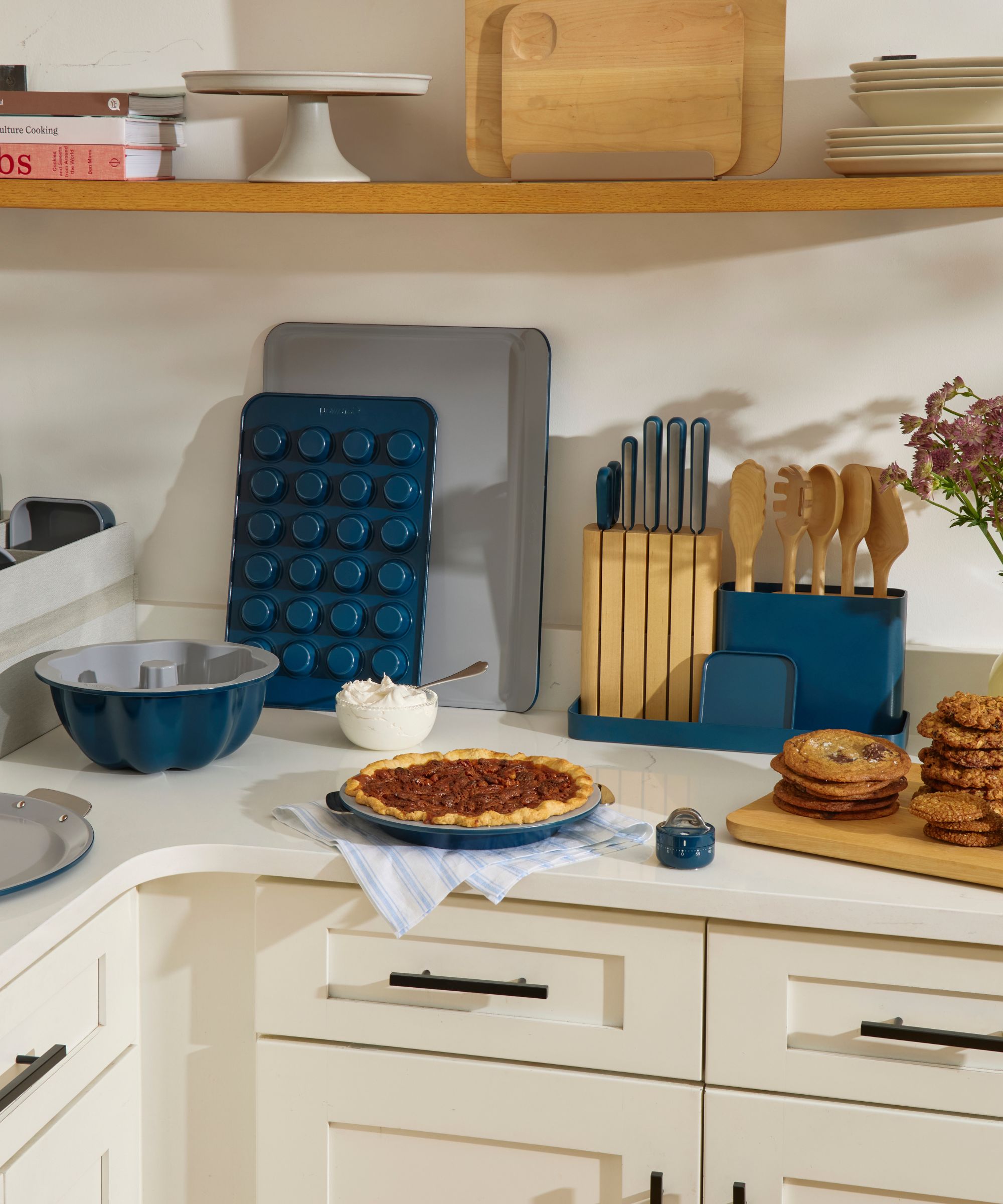 The blue Caraway bakeware+ range, including a muffin tin, a Bundt tin, and a pie tin on a white kitchen counter, displayed with wooden kitchen utensils, a cutting board full of cookies, and a blue striped dish towel.