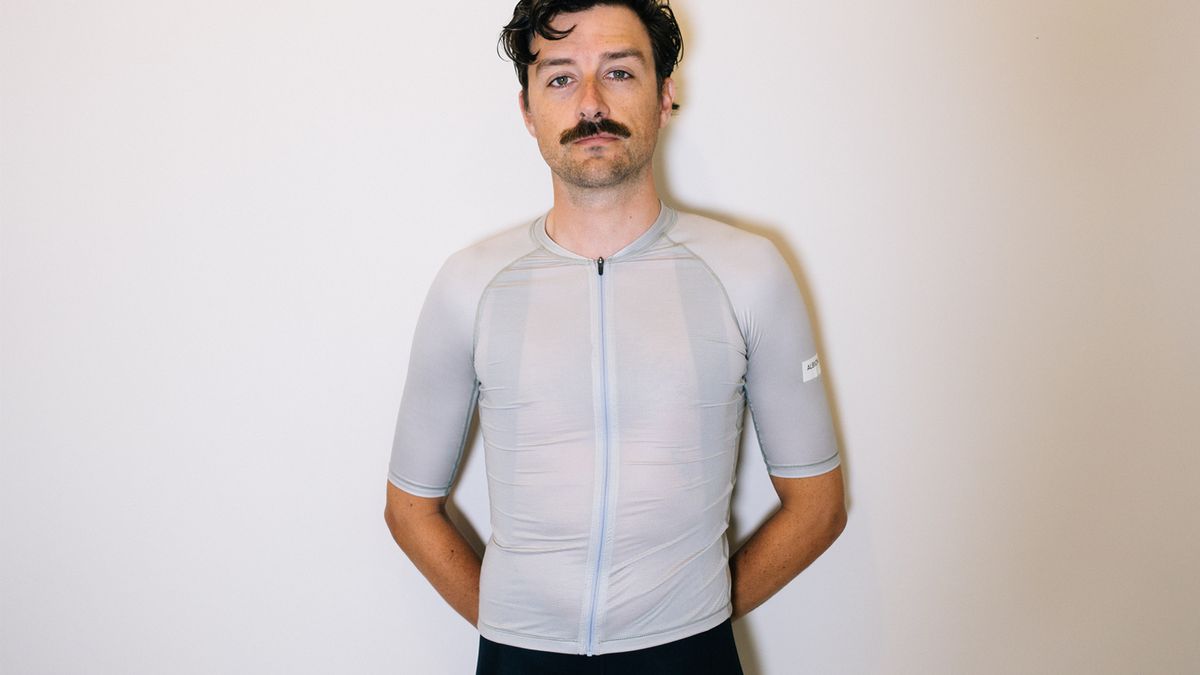 A white man stands against a white wall wearing black cycling shorts and a tight grey-green jersey