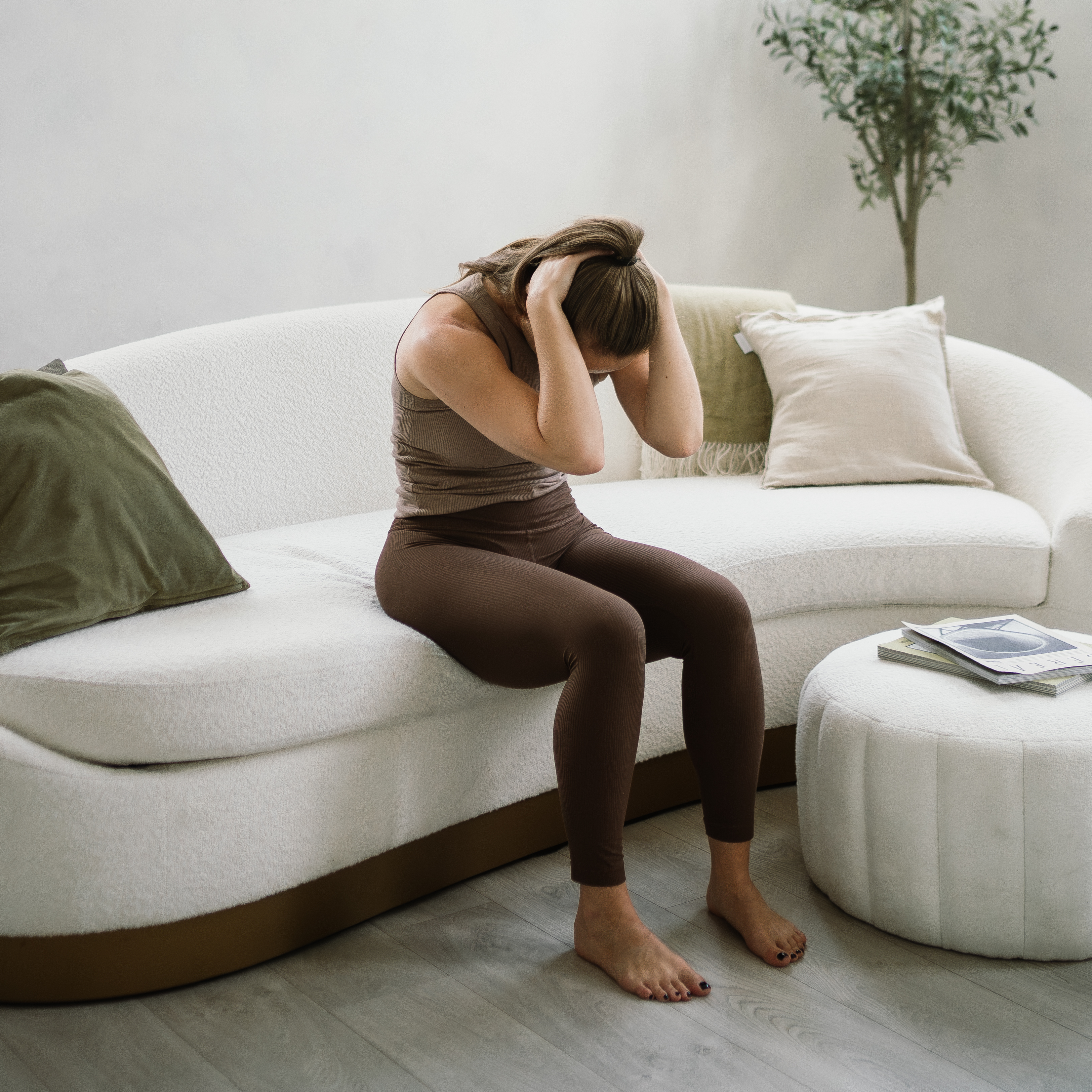 A woman sits on the edge of a curved white couch performing a spinal flexion stretch. Her knees are bent, feet flat on the floor, buttocks on the edge of the couch, and her hands are behind her head as she bends her spine forward.