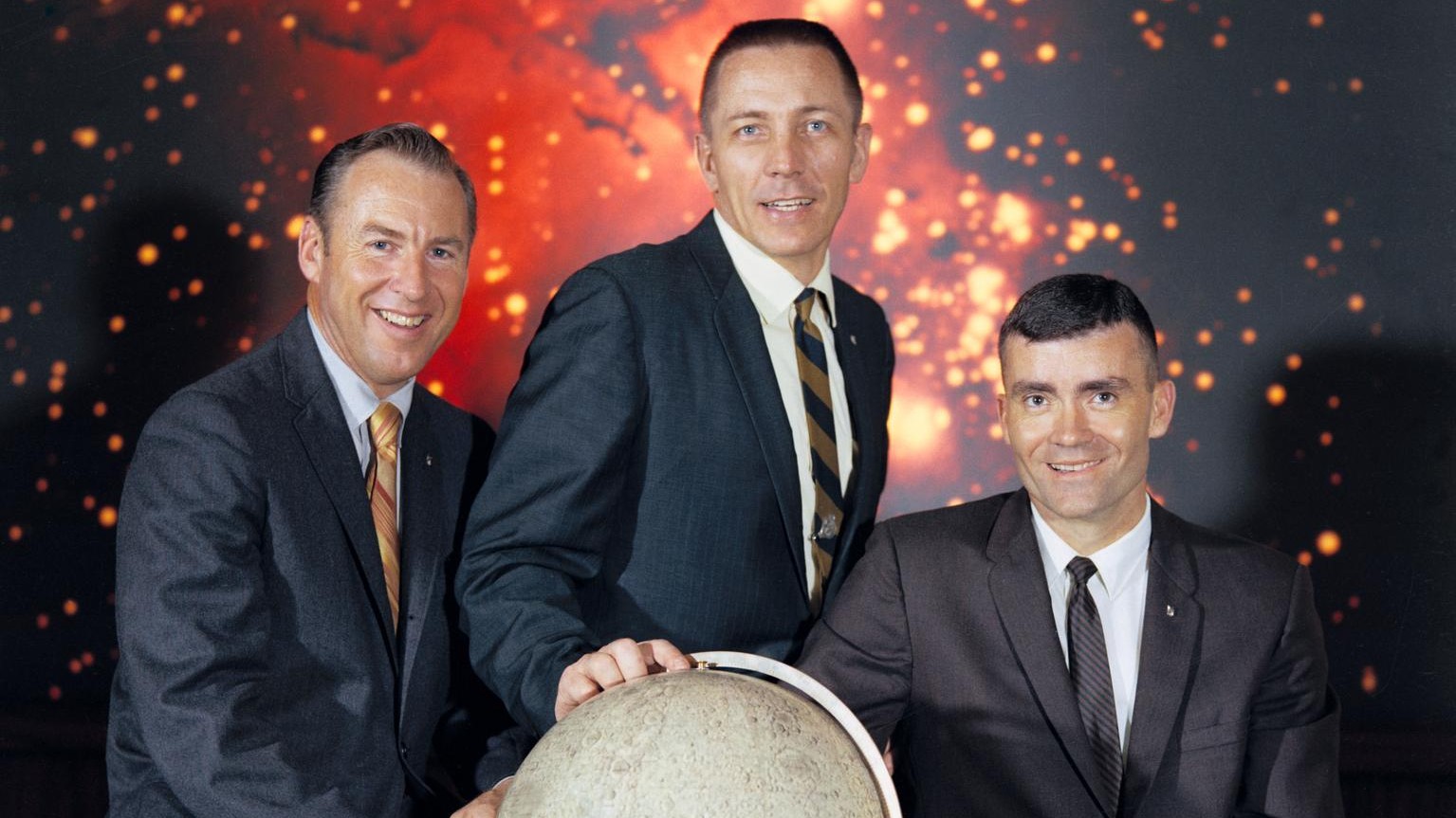three astronauts in suits stand in a row. the middle one, jack swigert, has his hand on a globe showing the moon. behind them is a picture of a nebula