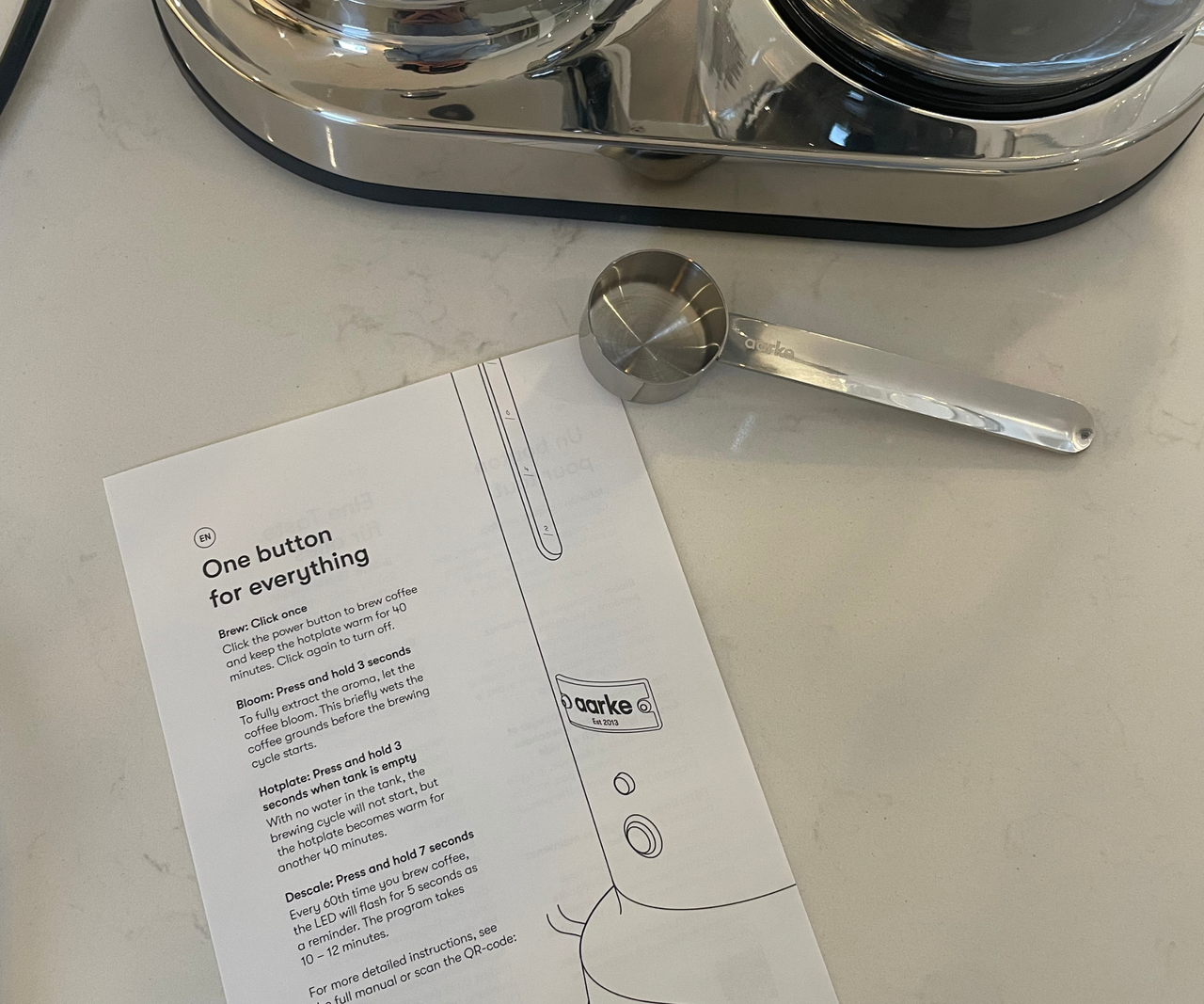 An instruction leaflet on a white kitchen counter next to a stainless steel coffee scoop.