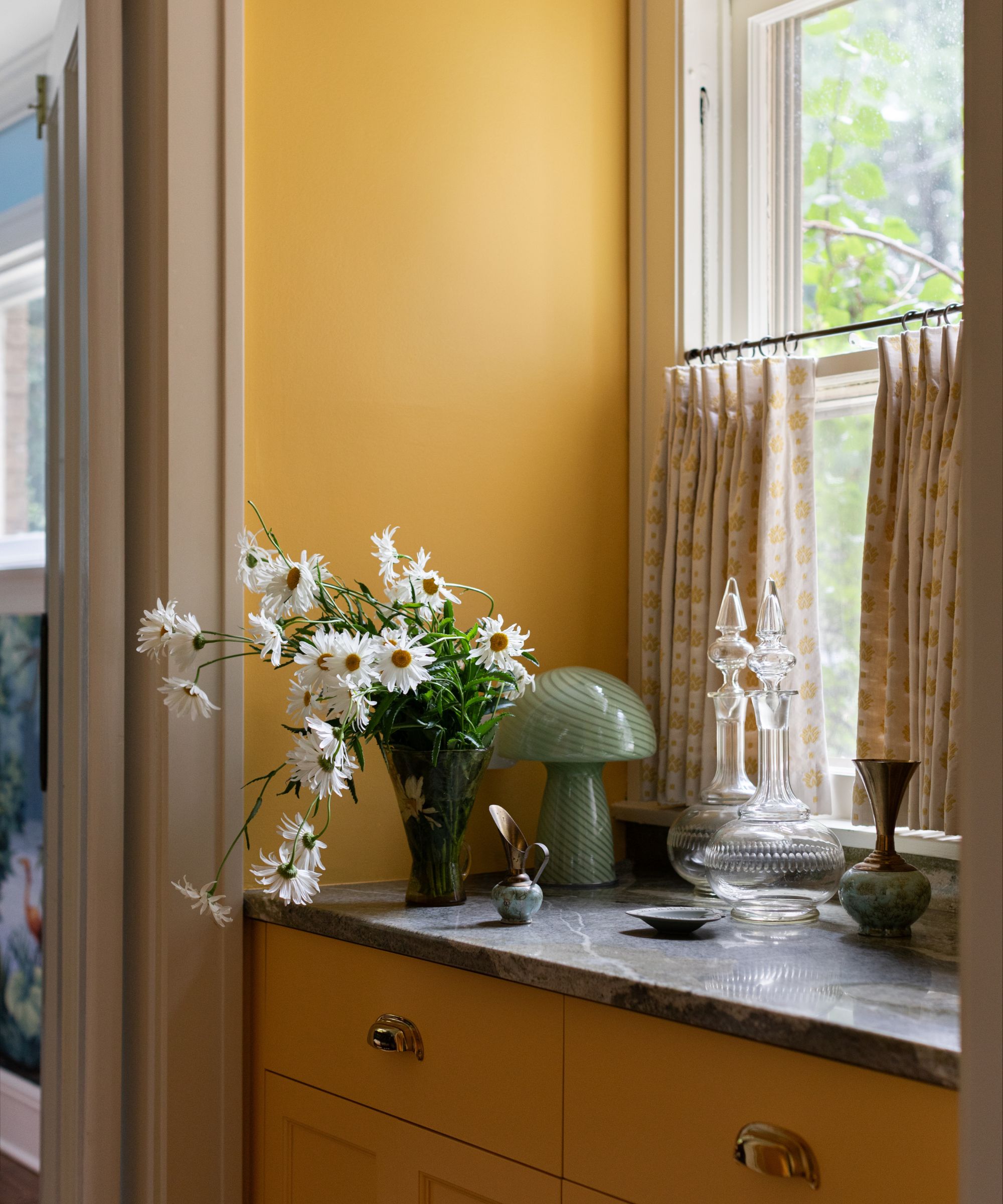 A corner of a bathroom with ochre walls and cabinets, cafe curtains on the window, and black countertops with a vase of white flowers and decorative items, and a lamp.