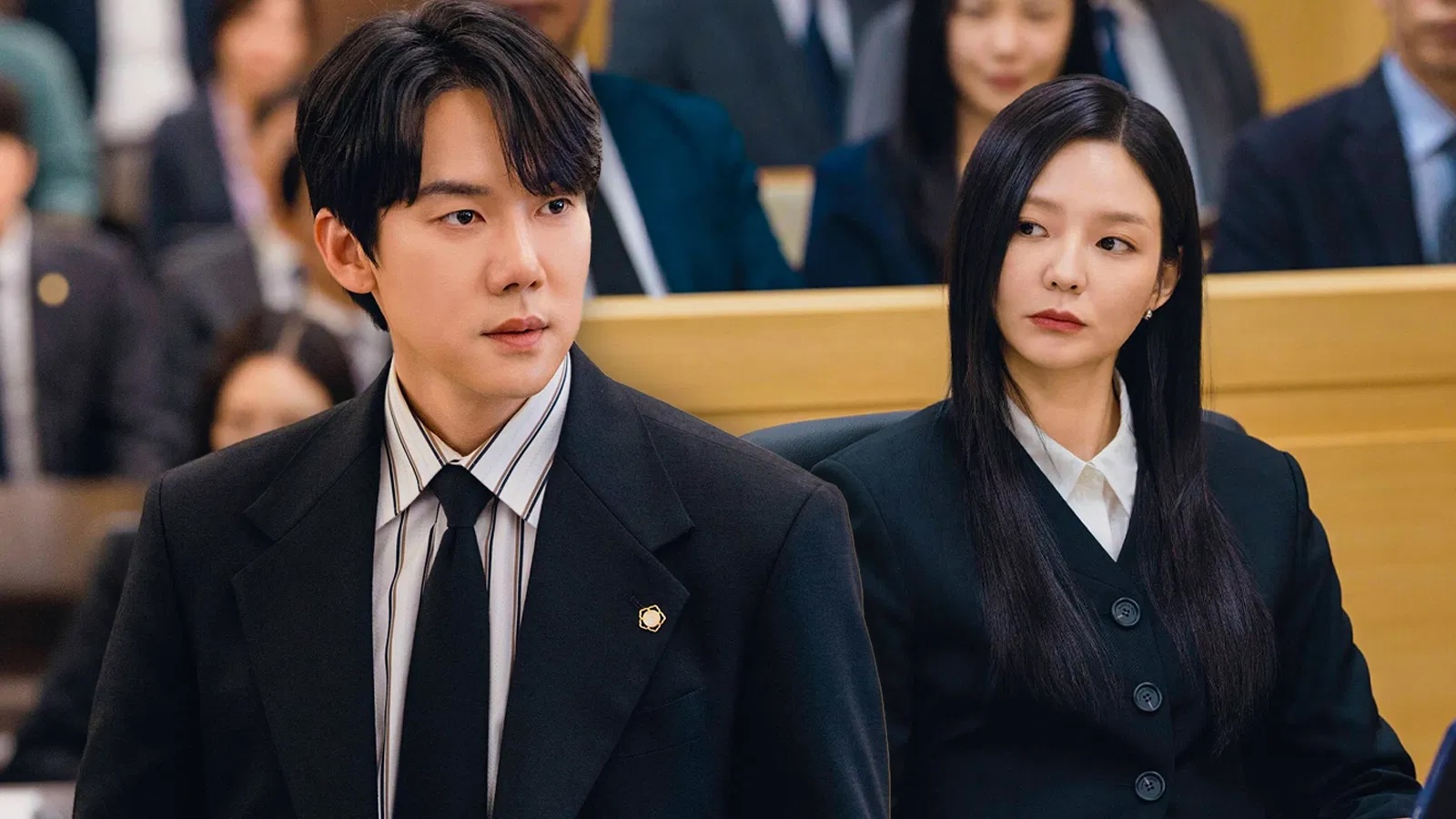 A man (Yoo Yeon-seok) and a woman (Esom) wear dark suits as they sit in a Korean courtroom in front of a crowded gallery, in the K-drama 'Phantom Lawyer.'