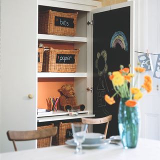 A storage cupboard next to a dining table with a blackboard on the door and labelled storage baksets on the shelves