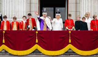 A balcony photo of King Charles's coronation featuring all of the page boys in red uniforms and The King and Queen smiling and waving in their crowns and robes