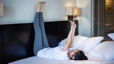 A woman practices yoga in bed. She is lying on her back, with her legs up against the headboard, pointing her foot to the ceiling. Her arms are stretched out and hands clasped together.
