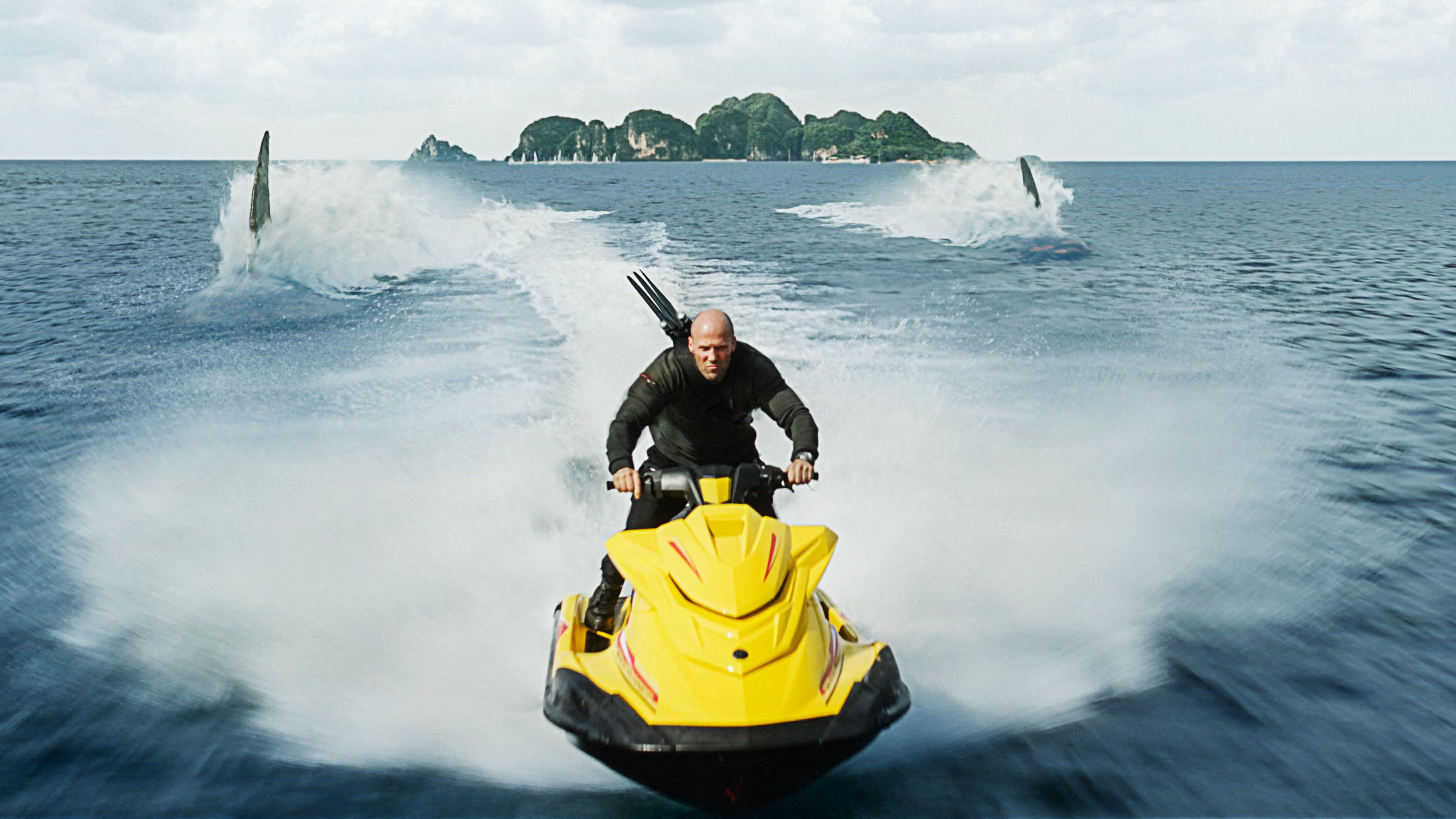 Jonas (Jason Statham) speeds away from two pursuing sharks on a yellow jet-ski in a scene from "Meg 2: The Trench"