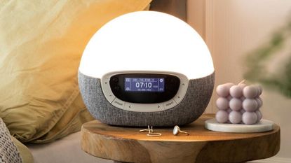 A Lumie Bodyclock Shine 300 sunrise alarm clock on a wooden bedside table next to a pastel lilac bubble cube candle