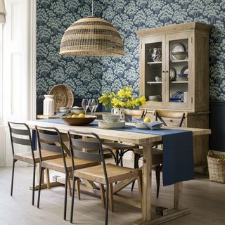 A dining room with wooden table, chairs, cupboard and pendant light with blue floral wallpaper