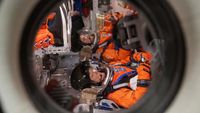 a look through a circular spacecraft window to see two astronauts in spacesuits lying on their backs 
