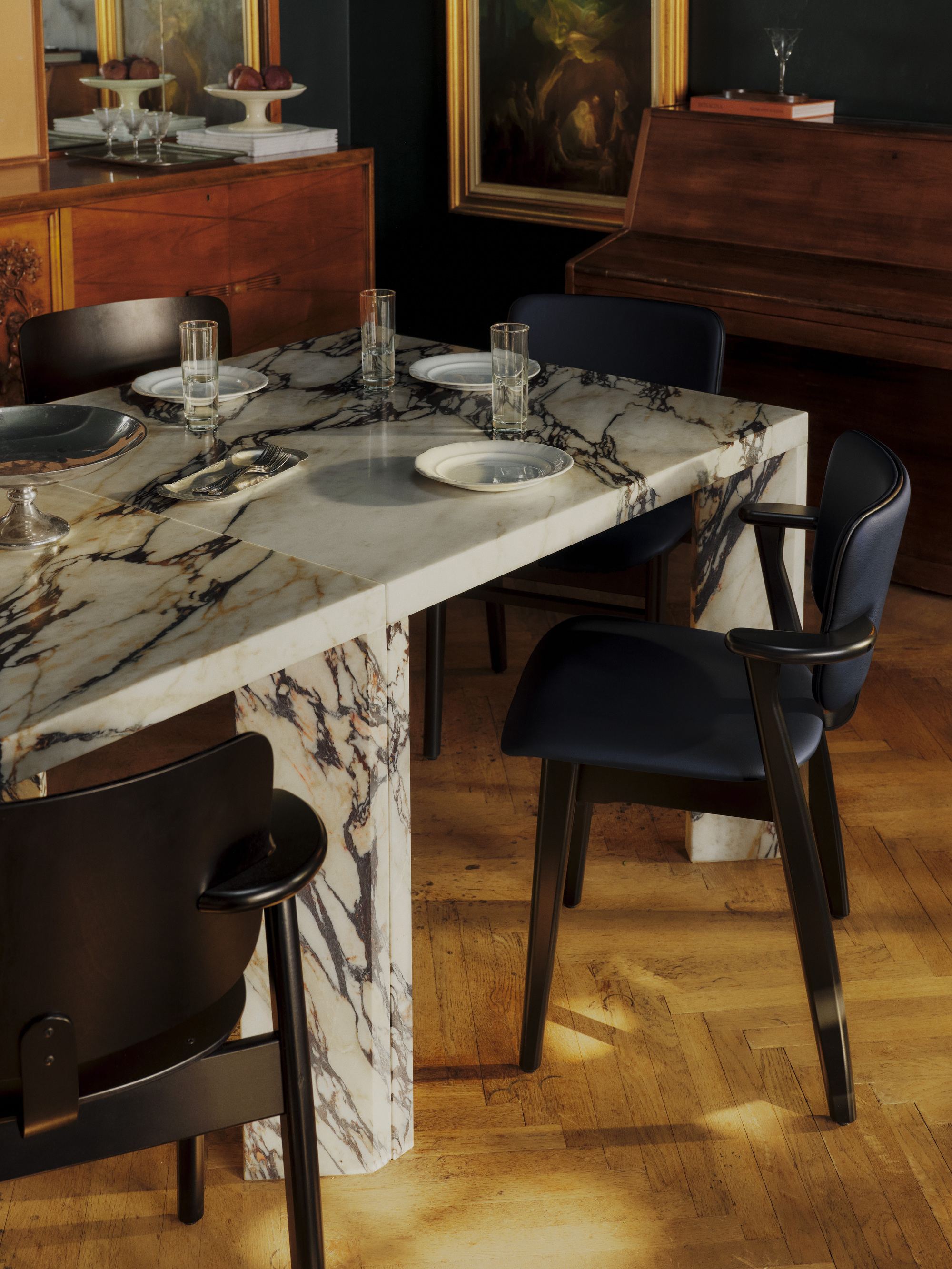 A navy blue leather and dark wood dining chair around a white and brown marble dining table. There are wooden floors, a wooden piano, and wooden cabinet.