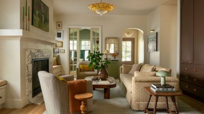 Warm neutral living room with a marble fireplace, off-white walls, cream patterned sofa, a striped armchair with orange throw, and glass doors leading into kitchen.
