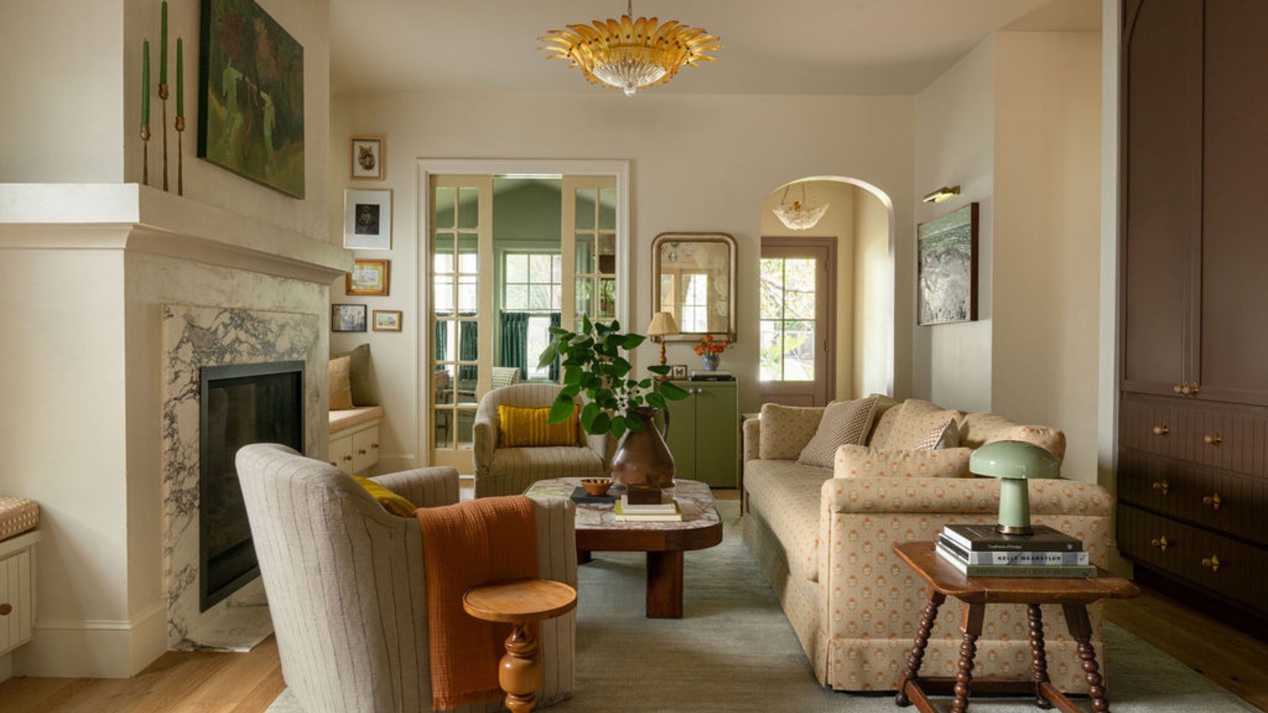 Warm neutral living room with a marble fireplace, off-white walls, cream patterned sofa, a striped armchair with orange throw, and glass doors leading into kitchen.