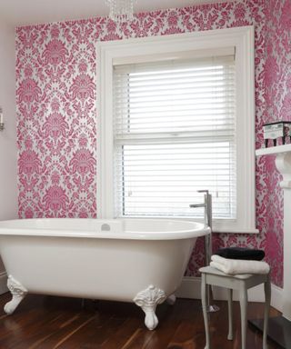 A bathroom with a pink and white ornate accent wall with a window, a white bathtub, a stool with towels on, and dark brown wooden floors