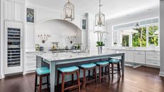 Marble kitchen island ideas are so chic. Here is a large marble kitchen island with five bright blue stools in front of it, two large gold pendant lights above it, and a countertop and two gold wall sconces behind it