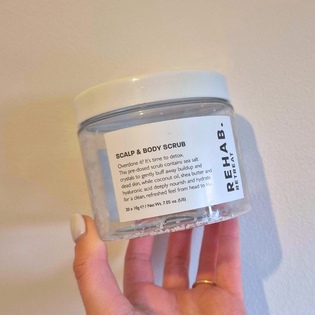 Hand holding empty tub of Rehab. Retreat Scalp &amp;amp; Body Scrub against white wall