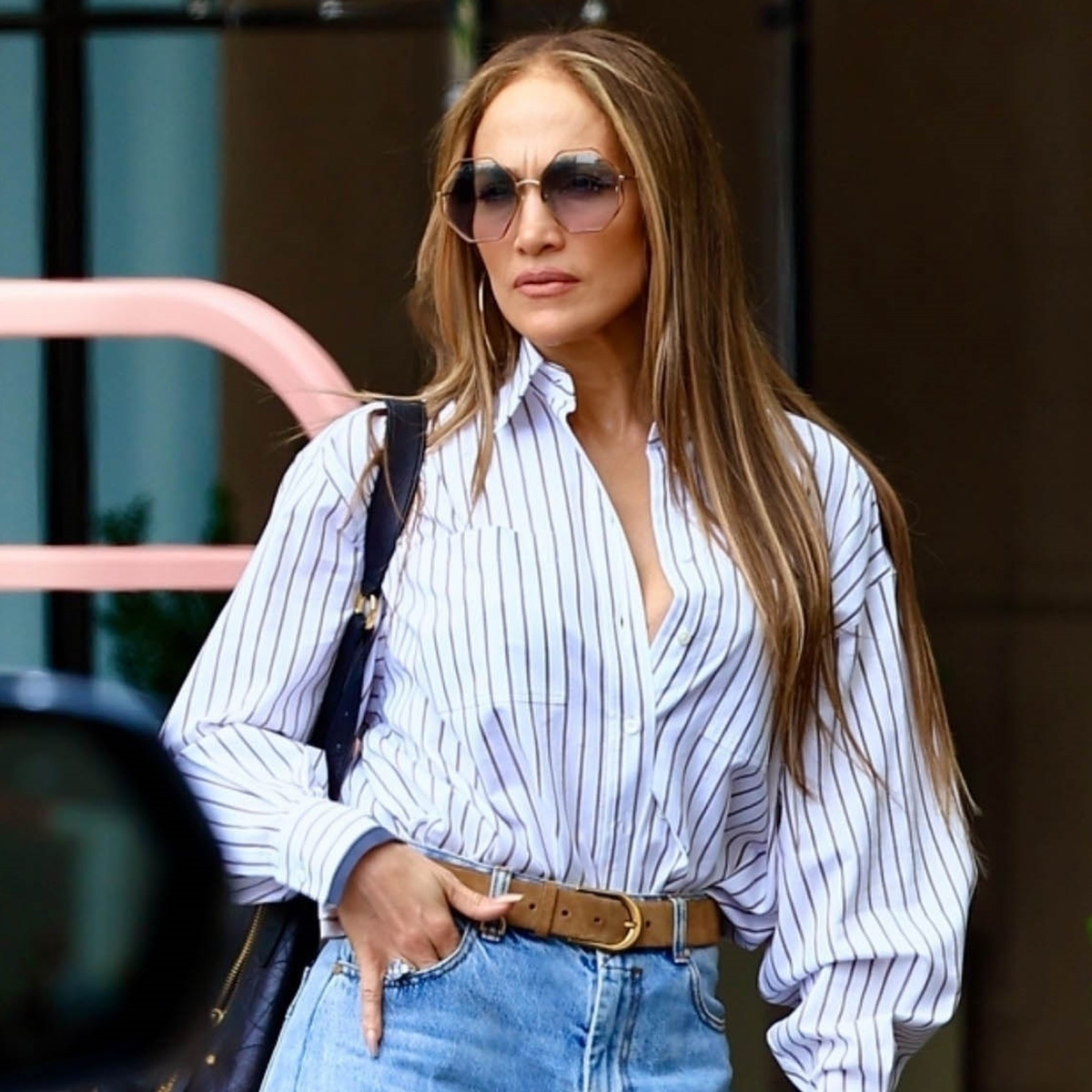 J.Lo wears a stripe button-down shirt, brown belt, wide-leg jeans, and a black shoulder bag.
