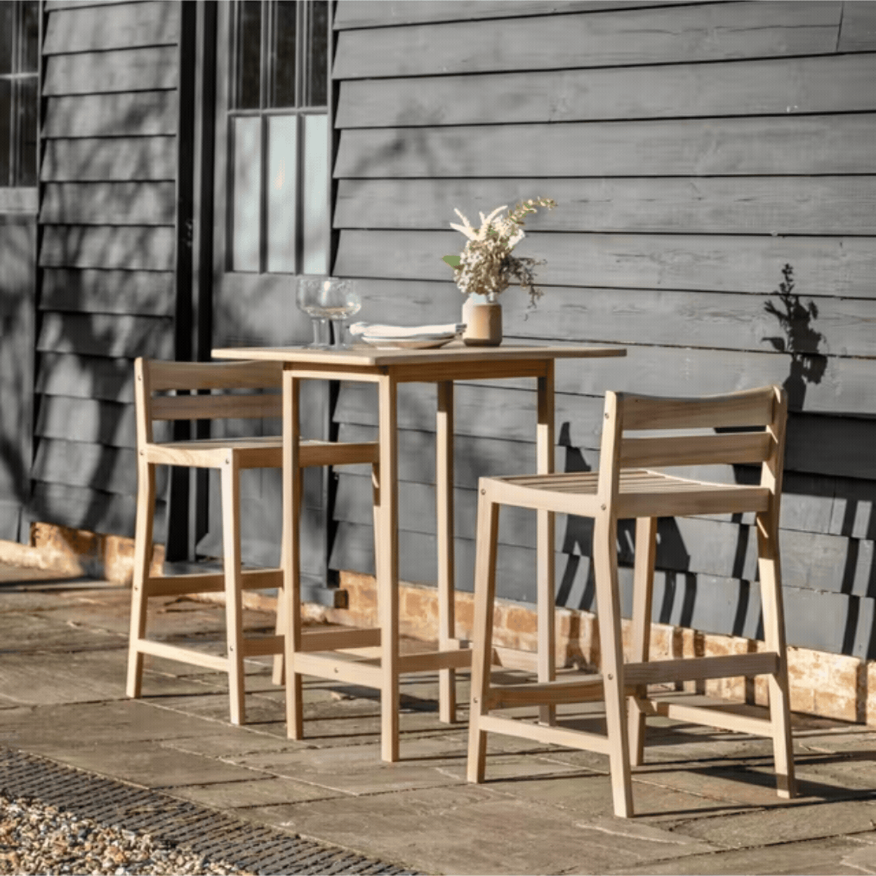 Astakos Acacia Wood 2-Seater Square High Bar Table &amp;amp; Chairs Set, Natural in a rotating gif showcasing it out in the sunshine and as a product against a pale green background.