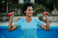 A woman stands outside holding a pair of light dumbbells in her hands at shoulder height and smiling.