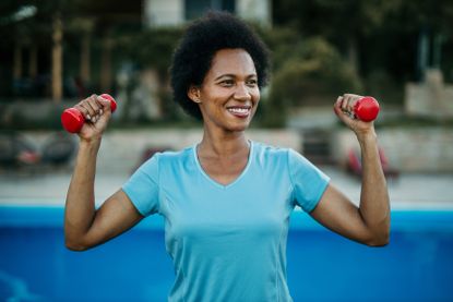 A woman stands outside holding a pair of light dumbbells in her hands at shoulder height and smiling.