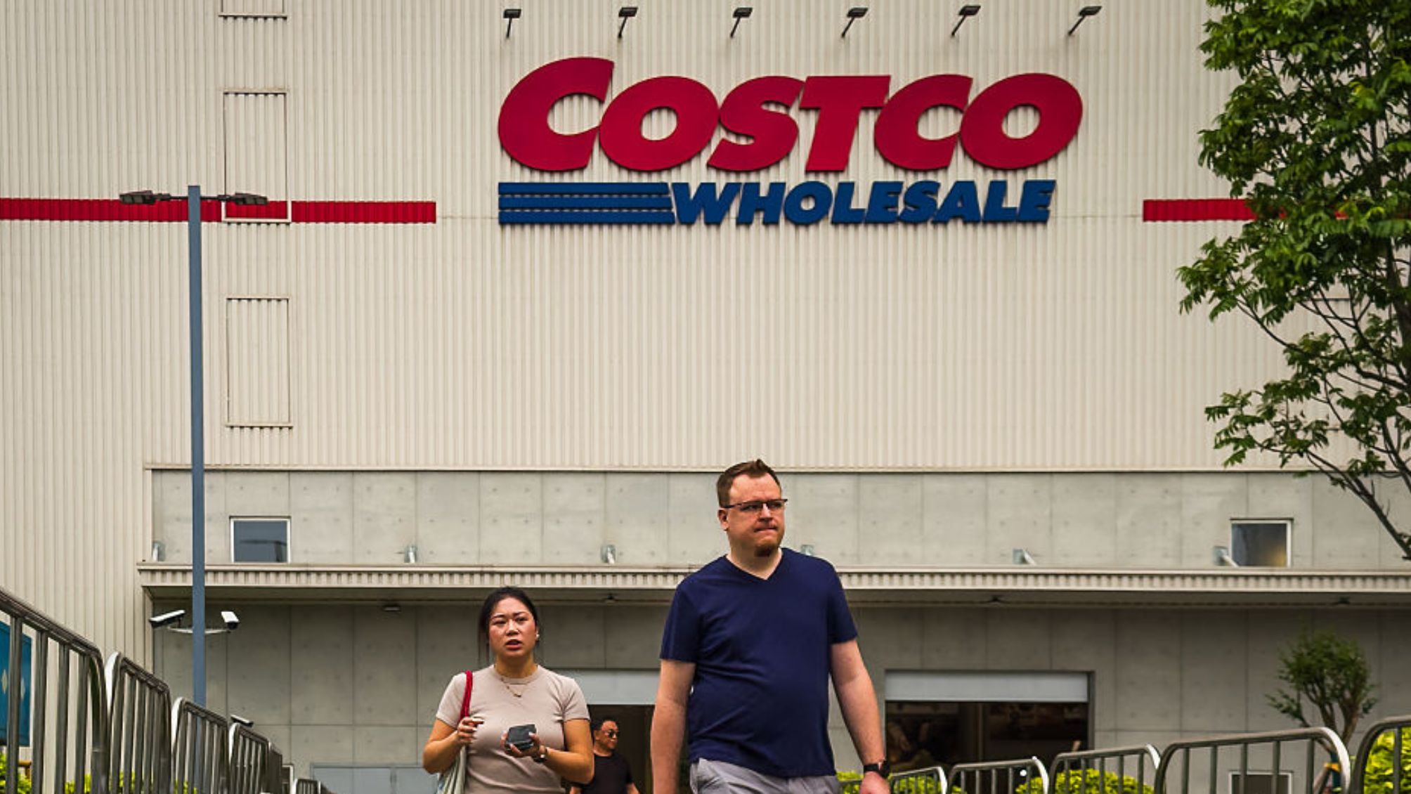 Shoppers walk along a fenced path outside a Costco Wholesale store, with the retailer's bold red and blue signage visible on the warehouse-style building