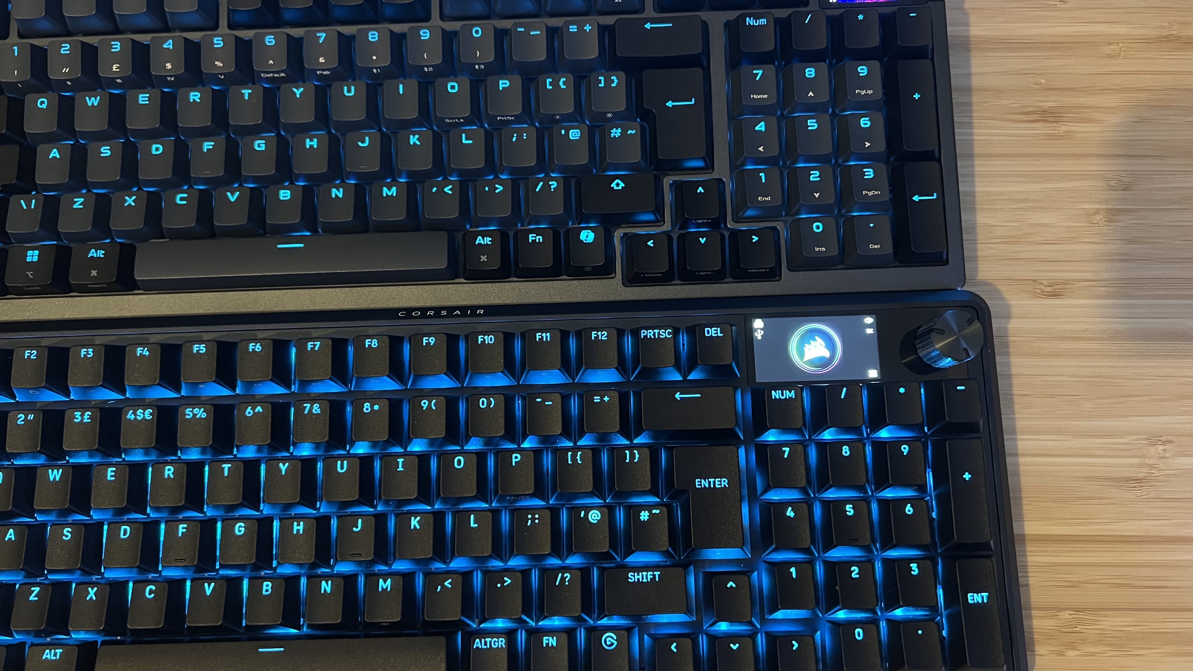 Asus ROG Azoth 96 HE and Corsair Vanguard Pro 96 gaming keyboards on a wooden desk next to each other, showing difference in key spacing
