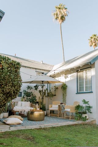 A corner backyard patio with woven furniture, cushions, and a blue rug next to a white house with a lawn in front of it