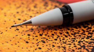 A close-up, shallow depth-of-field shot of the tip of a light gray mechanical pencil, a Winsor & Newton fineliner, with black accents, resting on a textured orange and black patterned surface.