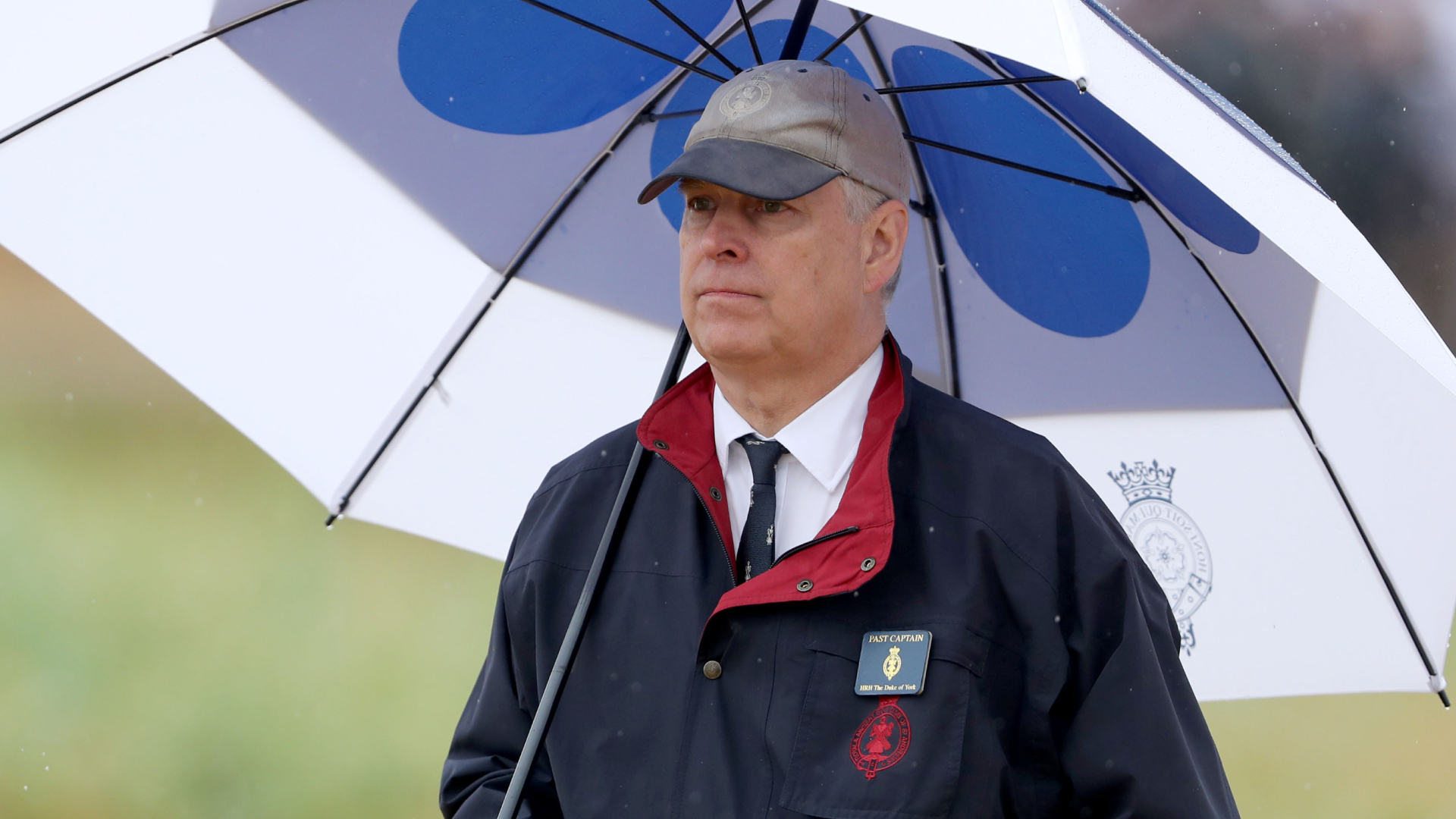 Prince Andrew stands under a white and navy umbrella in the rain while on the golf course, he wears a baseball cap and a navy jacket with a red trim