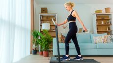 A woman walks on on a treadmill at home in her living room, wearing leggings, a sports bra and sneakers. Behind her is a couch, bookshelf and a few potted plants.