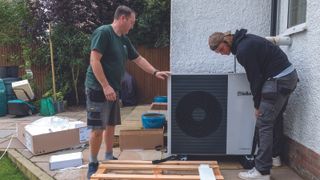 Two men installing an air source heat pump to the side of a home