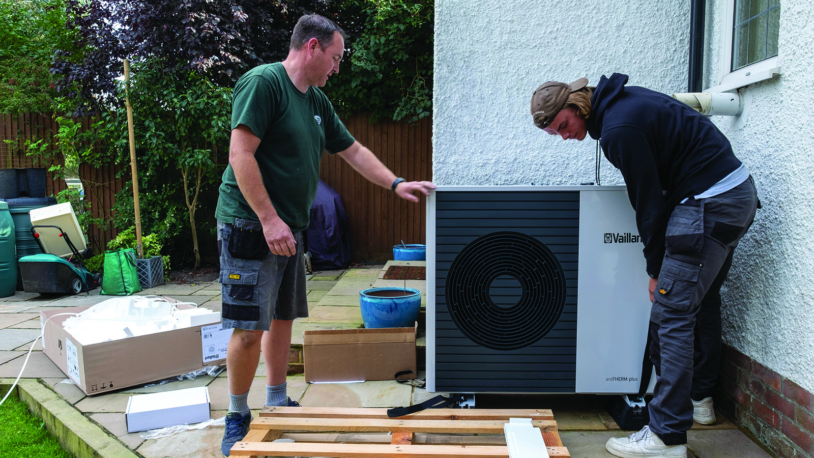 Two men installing an air source heat pump to the side of a home