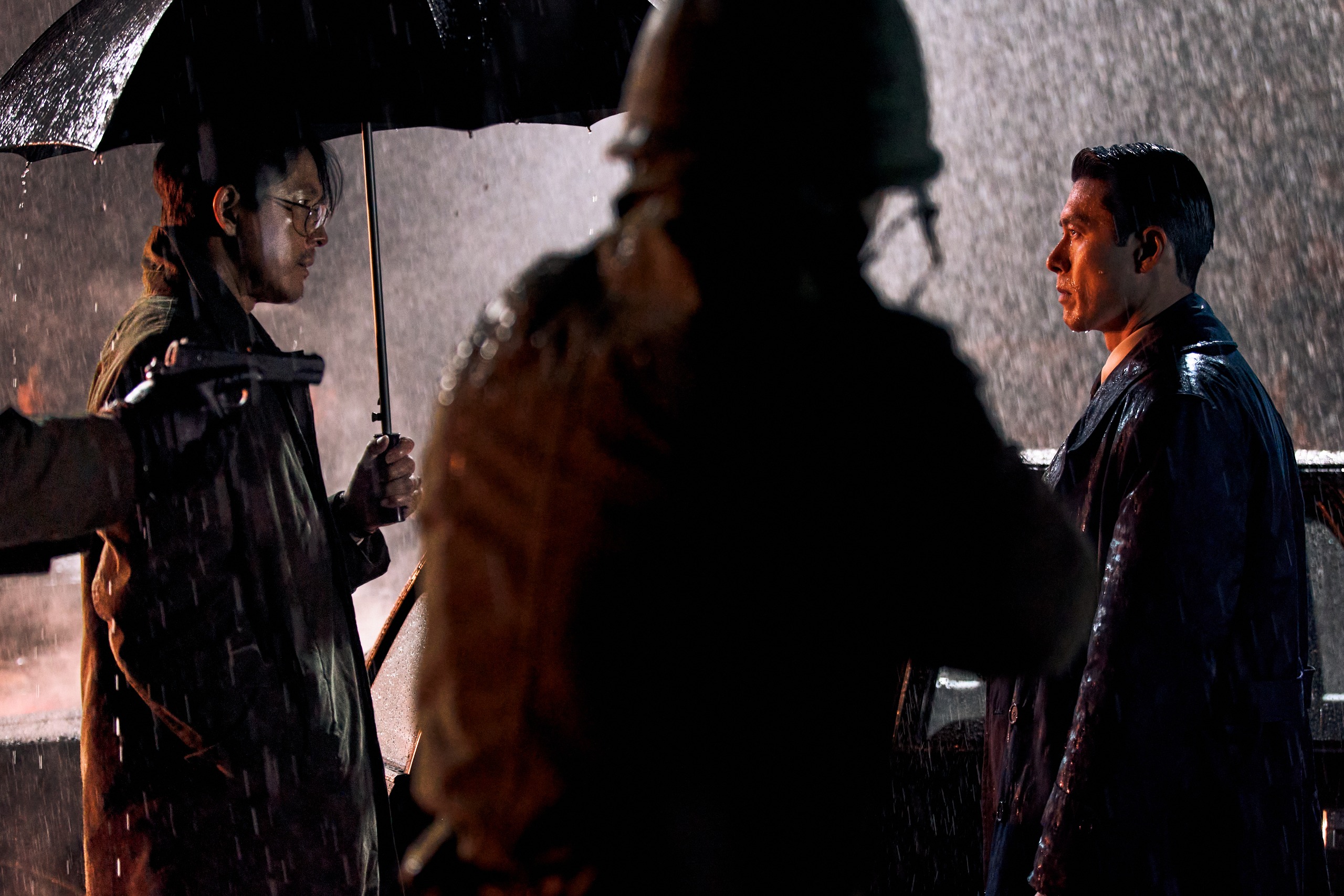 Two men look at each other while standing in the rain at night, as a military soldier's back is seen between them. The man on the left (Jung Woo-sung as Jang Geonyoung) wears glasses and holds an umbrella, while the man on the right (Hyun Bin as Baek Kitae) is uncovered. A still from Hulu's 'Made in Korea' season 2.