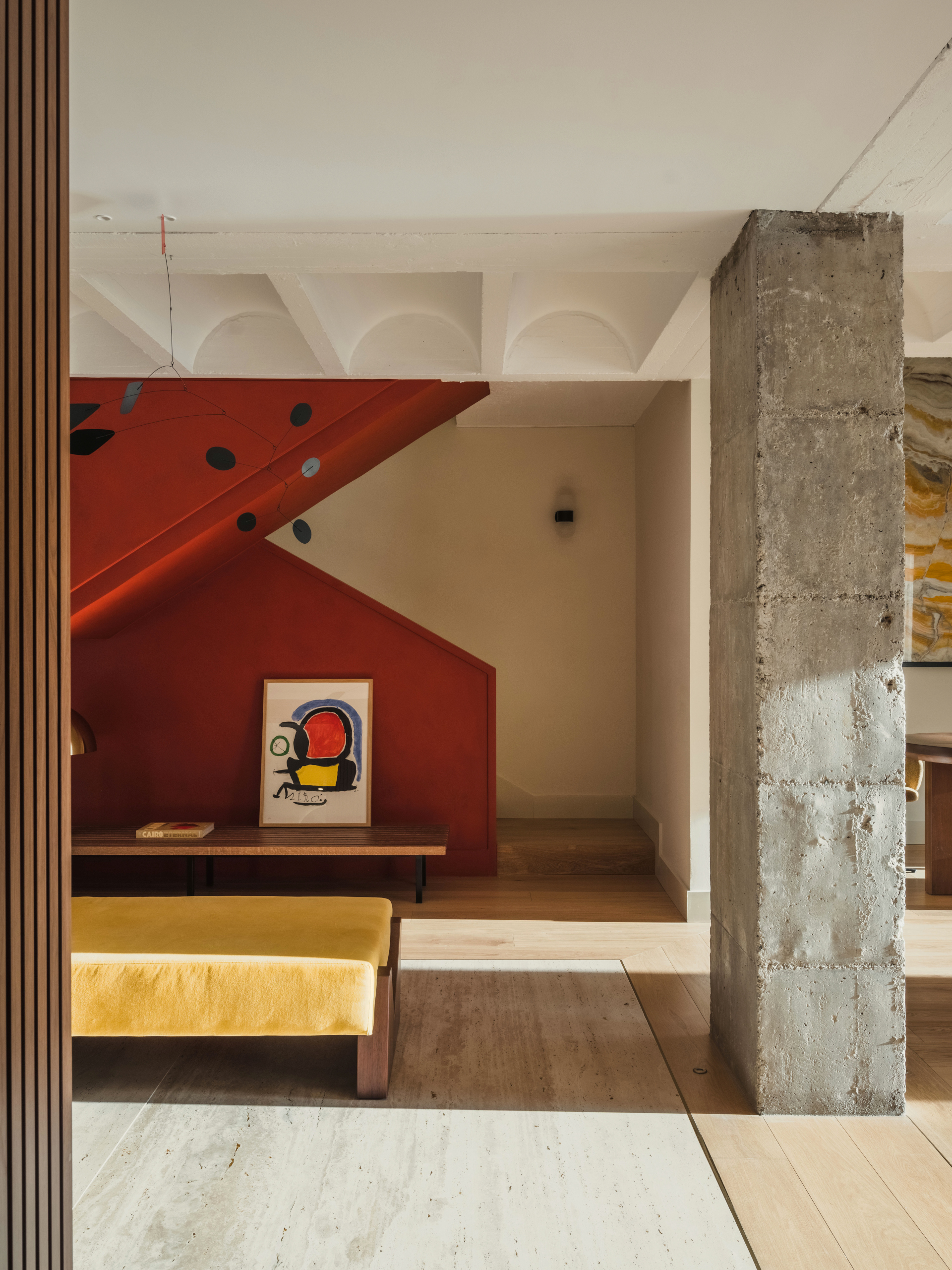A living room with white walls, concrete pillars, a mustard bench, and a red enclosed staircase that zig-zags in the background