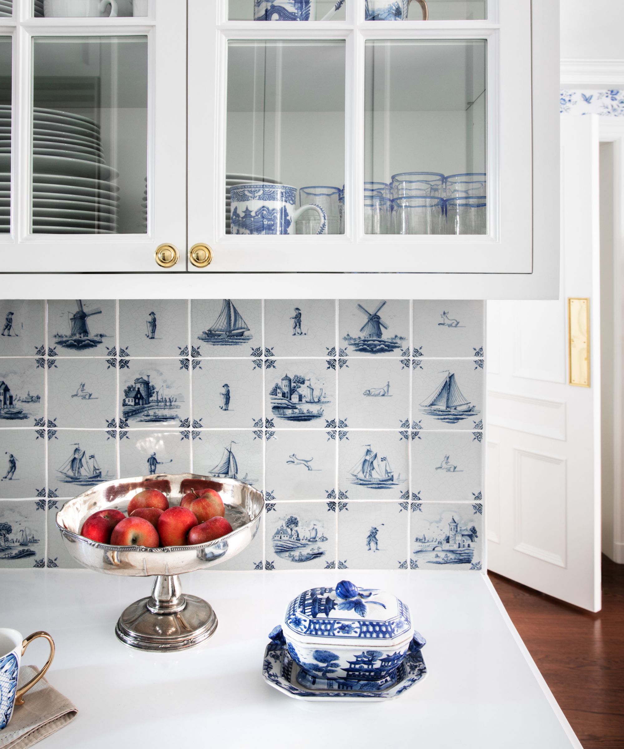 A kitchen corner showing white countertops, white class-fronted upper cabinets, and a Delft tile backsplash
