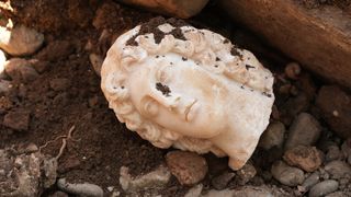A white marble bust of Alexander the Great, severed at the neck, atop dirt and rubble.