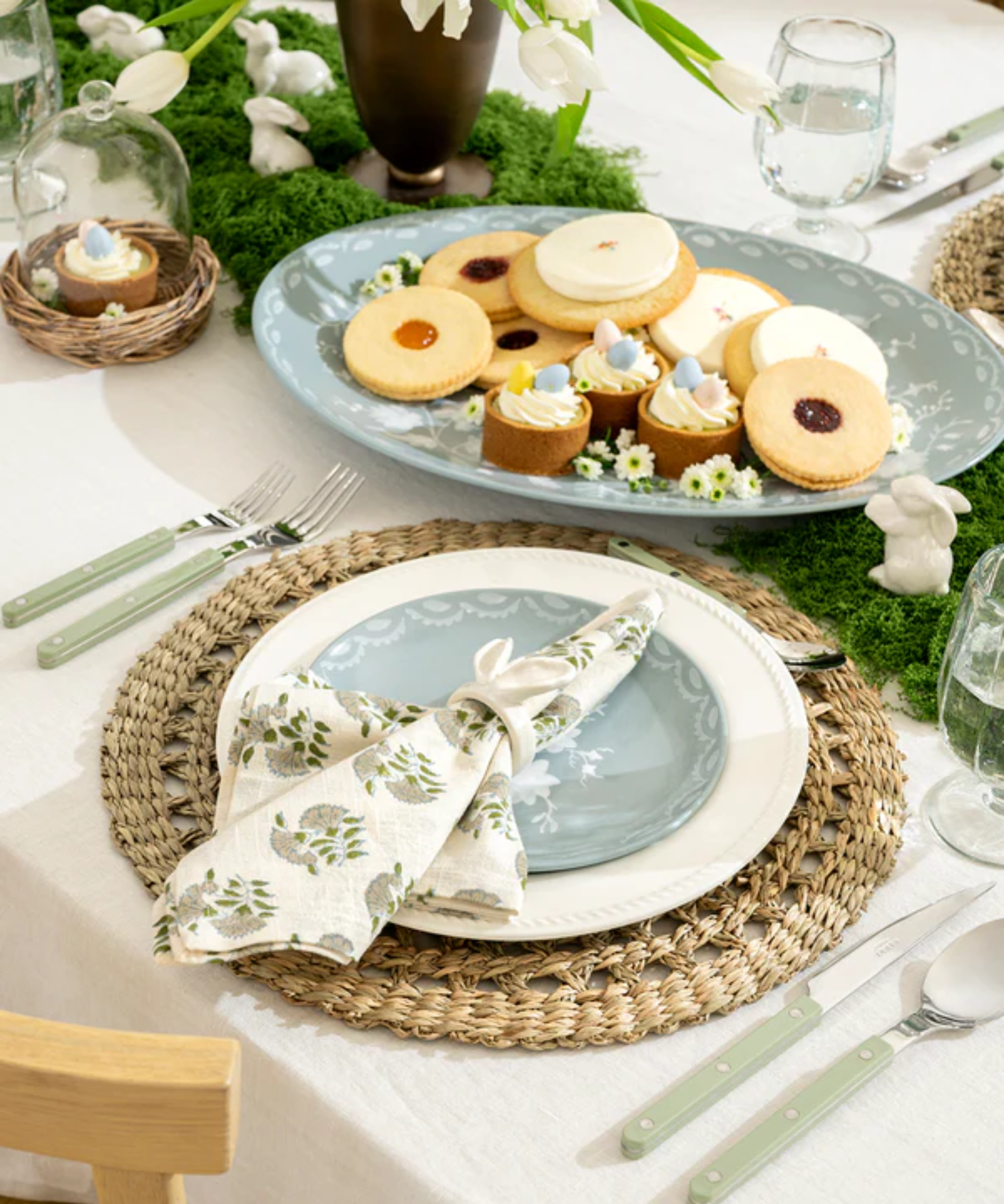 an Easter tablescape with a charger plate, a dinner plate, and a blue salad plate, a floral napkin, green cutlery, and a platter with cookies on it